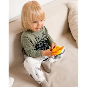 Little child with blond hair sits on a beige couch, holding an orange bird-shaped toy.