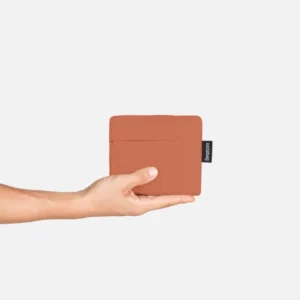 Hand presenting a small brown square wallet with a dark tag on the side against a light background.