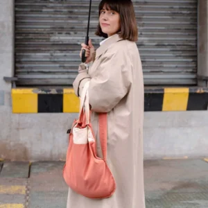 Woman in a beige trench coat holds a black umbrella while carrying a pink and cream tote bag in an urban setting with a closed shutter behind her.