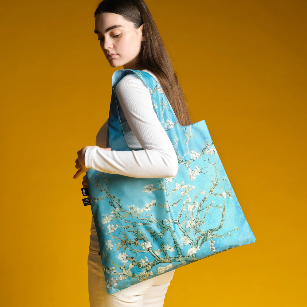 Woman walking left with a light blue tote bag featuring white blossom design over a mustard background.
