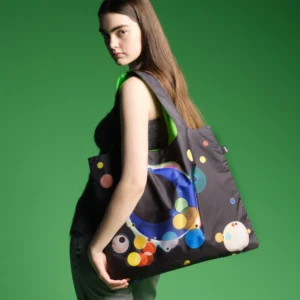 Side profile of a young woman carrying a large black tote bag with colorful circular patterns against a green background.