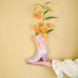 Pastel pink and lavender boot-shaped vase filled with orange flowers, held against a pale yellow wall by a gloved hand.